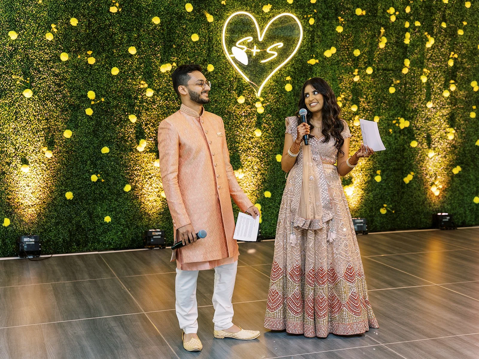 Couple in peach and gold traditional outfits holding microphones during engagement party speeches.