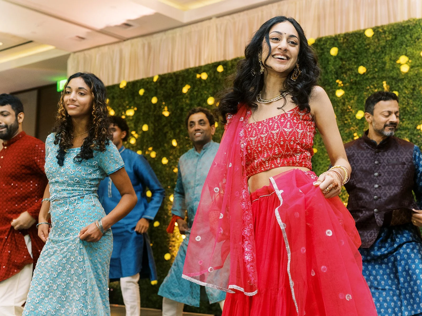 Women in red and blue lehengas dancing in front of green floral wall at wedding event.