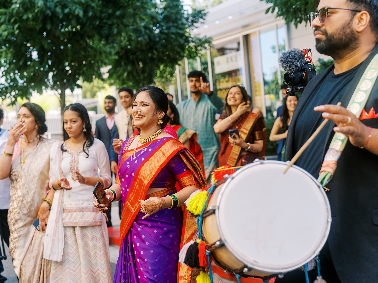 Guests dancing in colorful traditional outfits beside dhol player during outdoor Indian wedding procession.