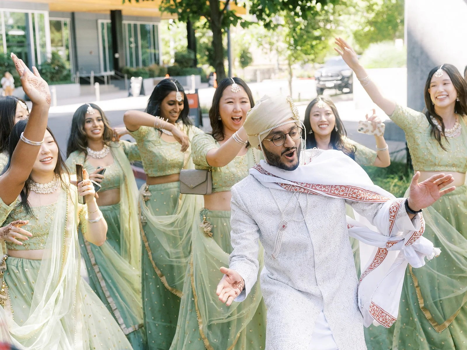Groom in white sherwani dancing in the street surrounded by bridesmaids in sage green lehengas.