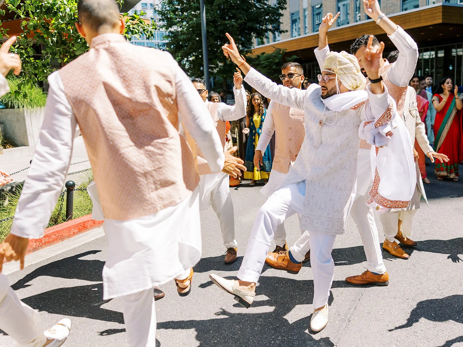 Groom in cream turban dancing with groomsmen in coordinated vests during daytime wedding procession.