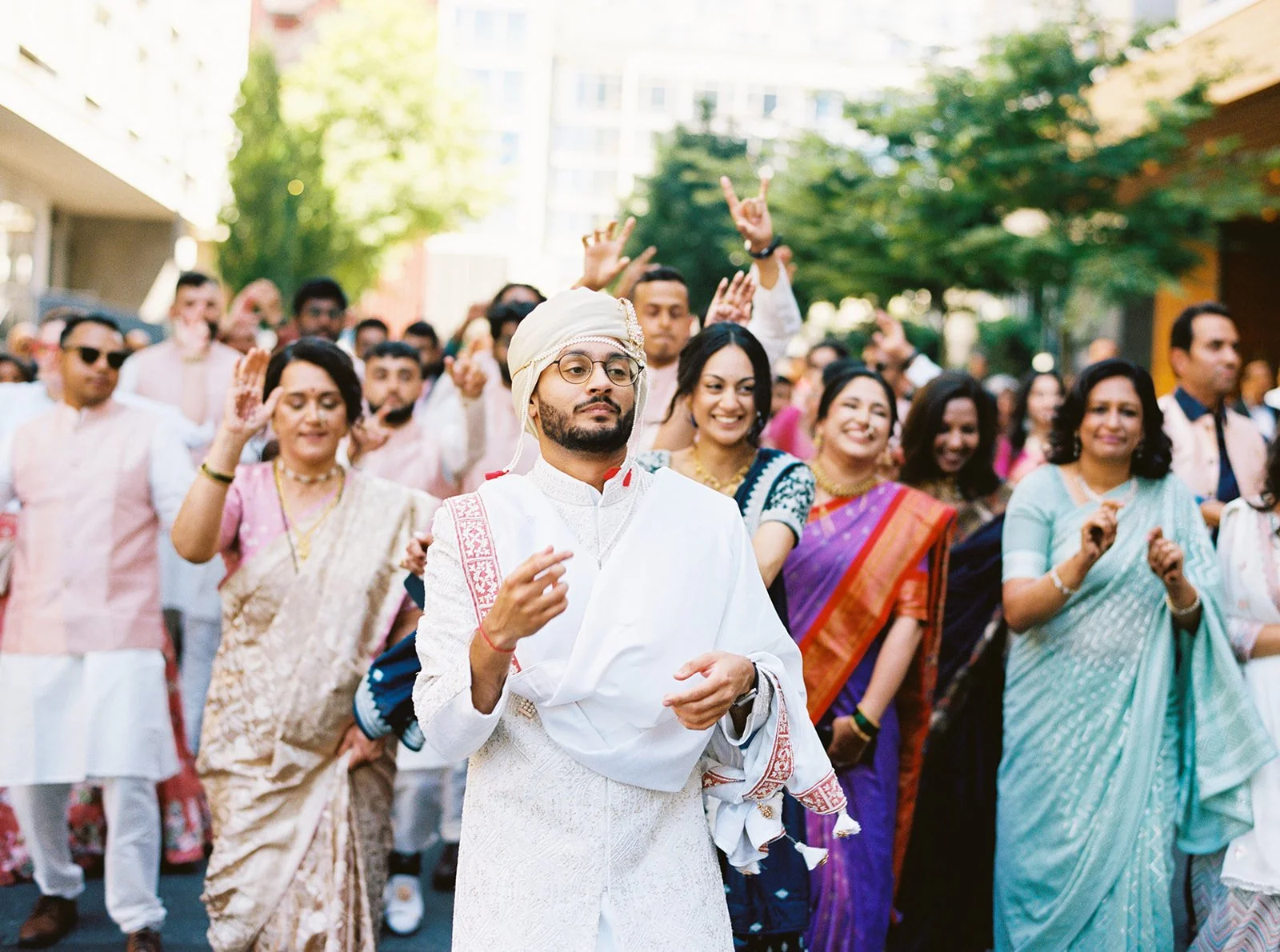Groom walking confidently at the front of baraat as family and friends dance behind him.