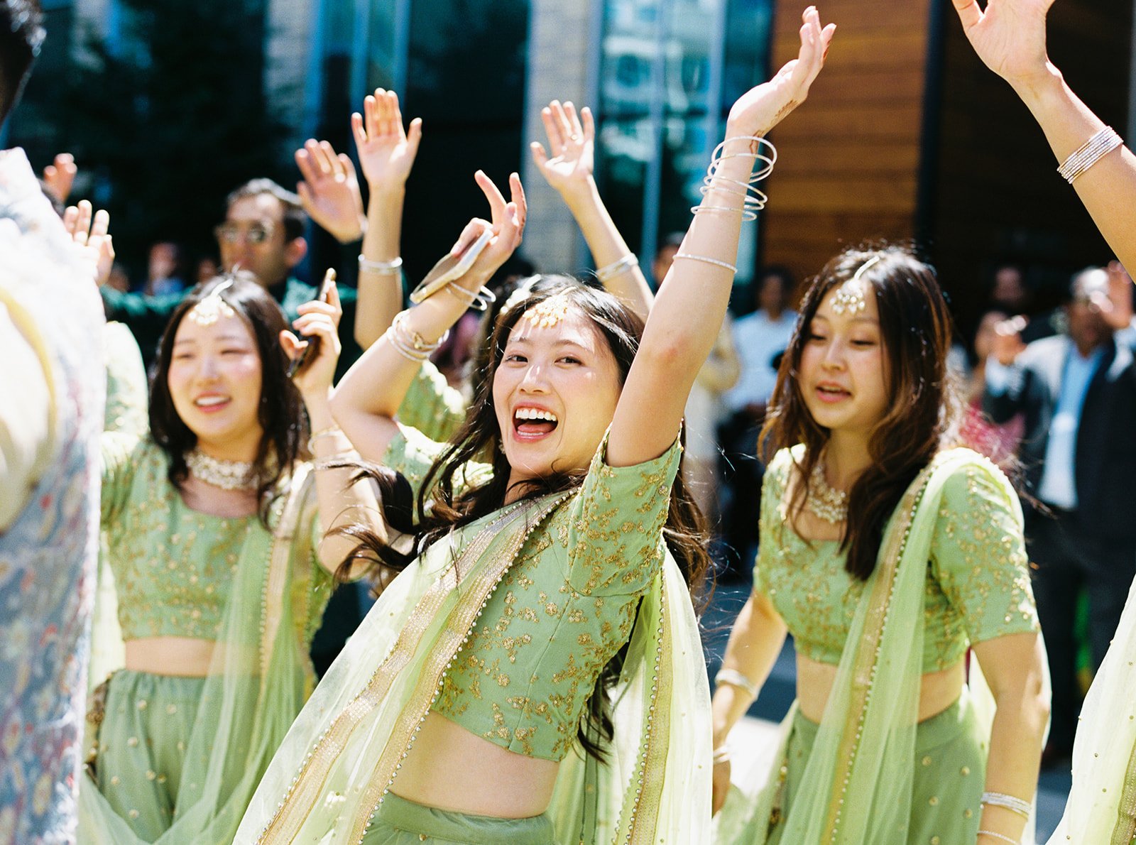 Bridesmaids in matching green lehengas raising their hands and dancing during outdoor wedding procession.