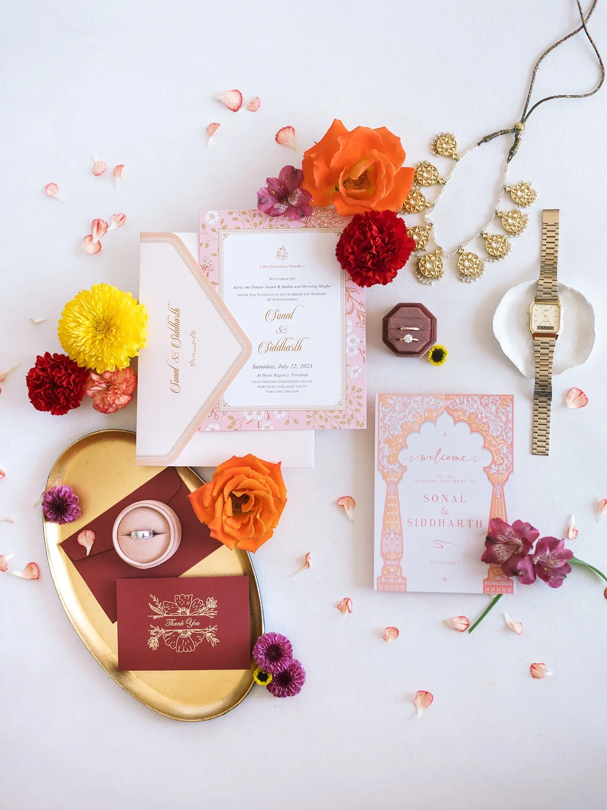 Flat lay of Indian wedding invitation suite with rings, gold jewelry, watch, and vibrant flowers.