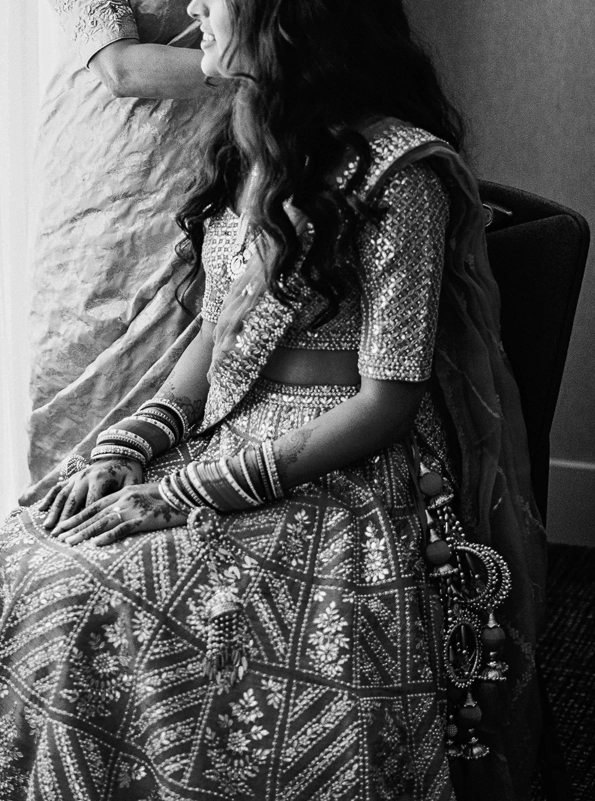 Bride seated by window in embroidered lehenga, hands with mehndi resting on her lap.