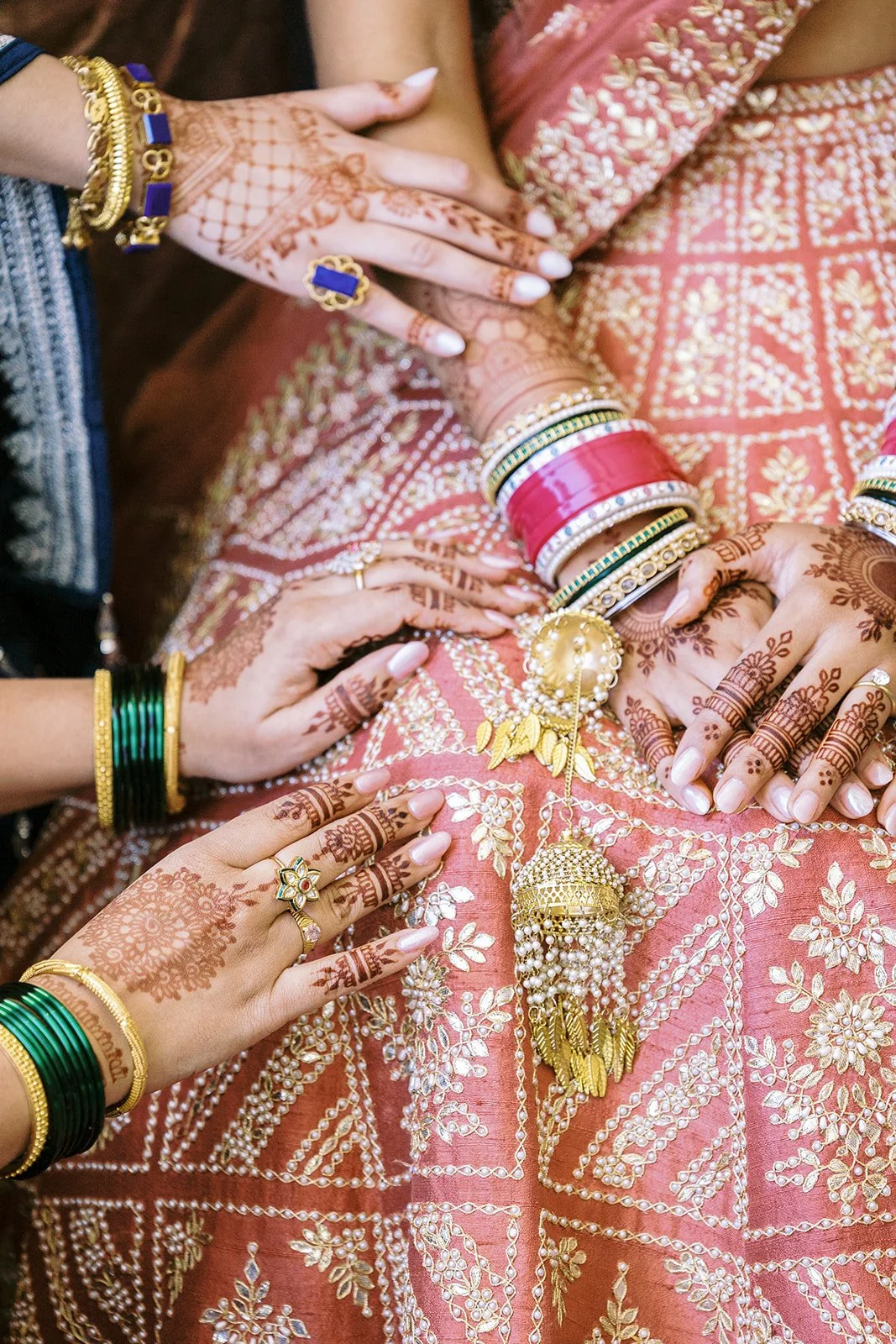 Close-up of bride’s henna hands and red chooda bangles surrounded by bridesmaids’ hands.