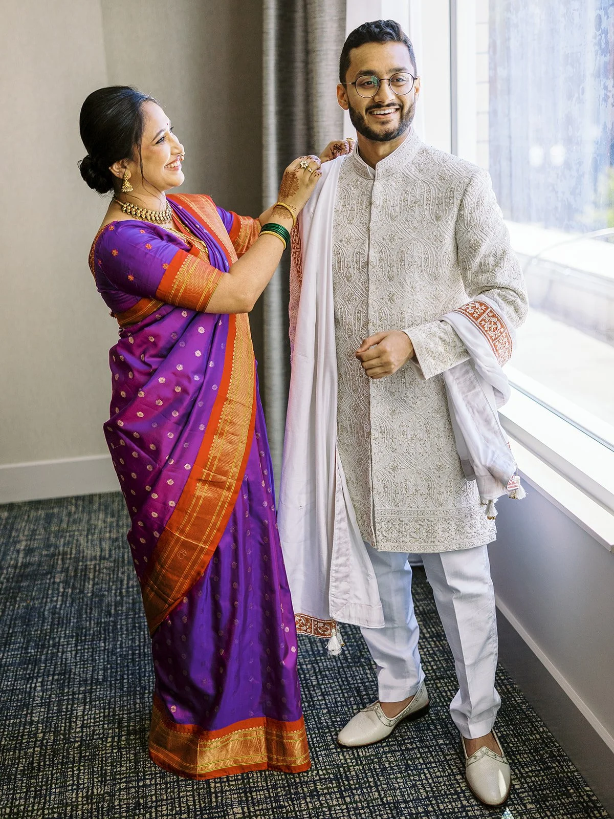 A woman in a purple saree helps a groom in an ivory sherwani adjust his dupatta during wedding preparations.