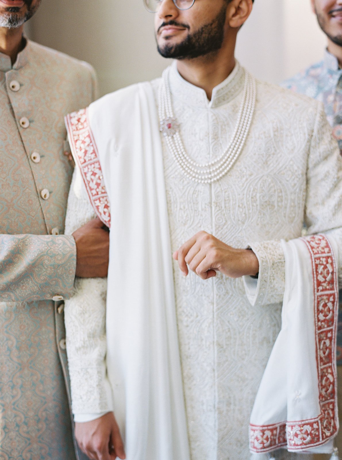A groom in a white embroidered sherwani with a pearl necklace, flanked by two men in traditional attire