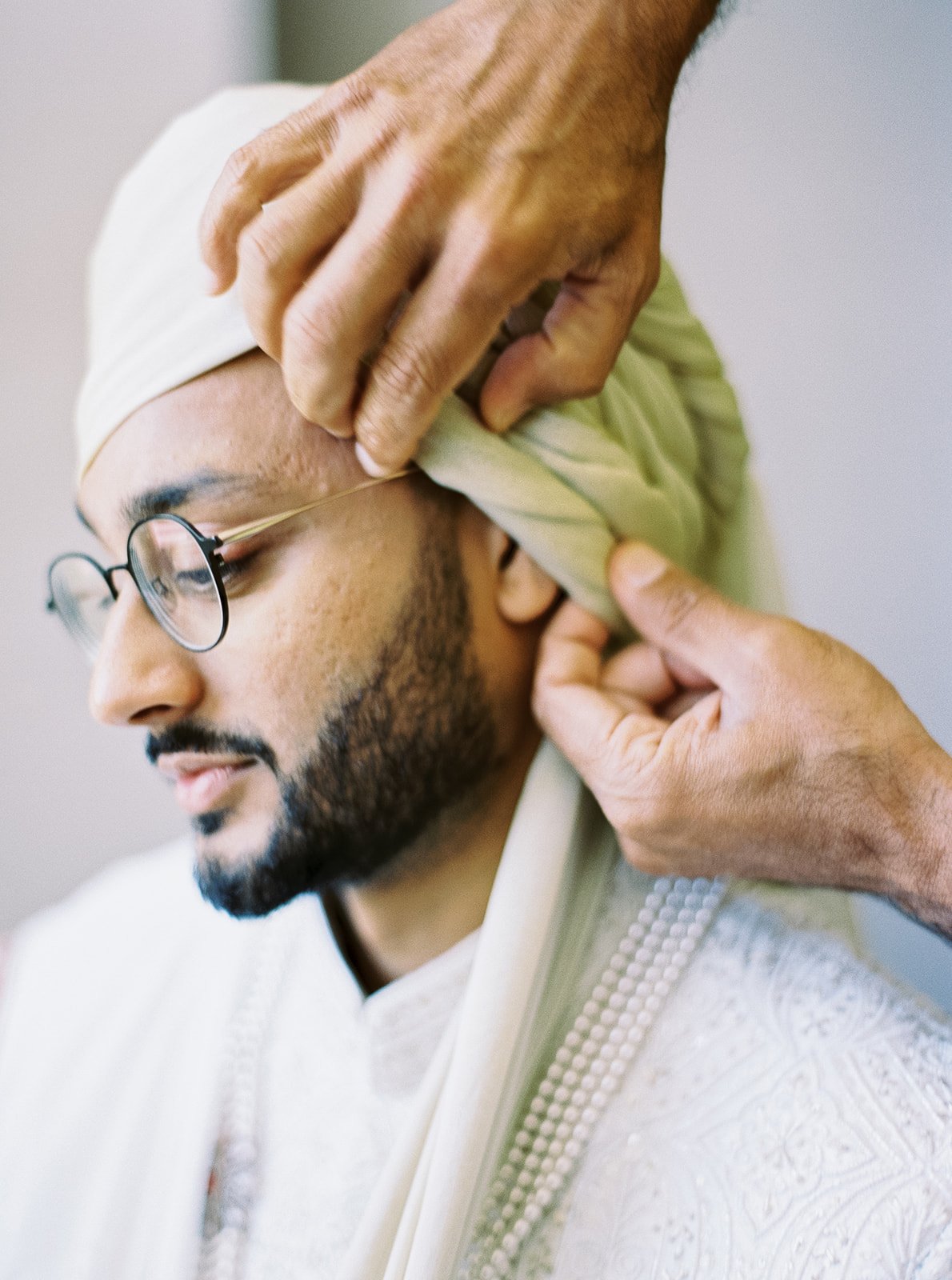 Close-up of a groom in glasses having his turban wrapped by unseen hands.