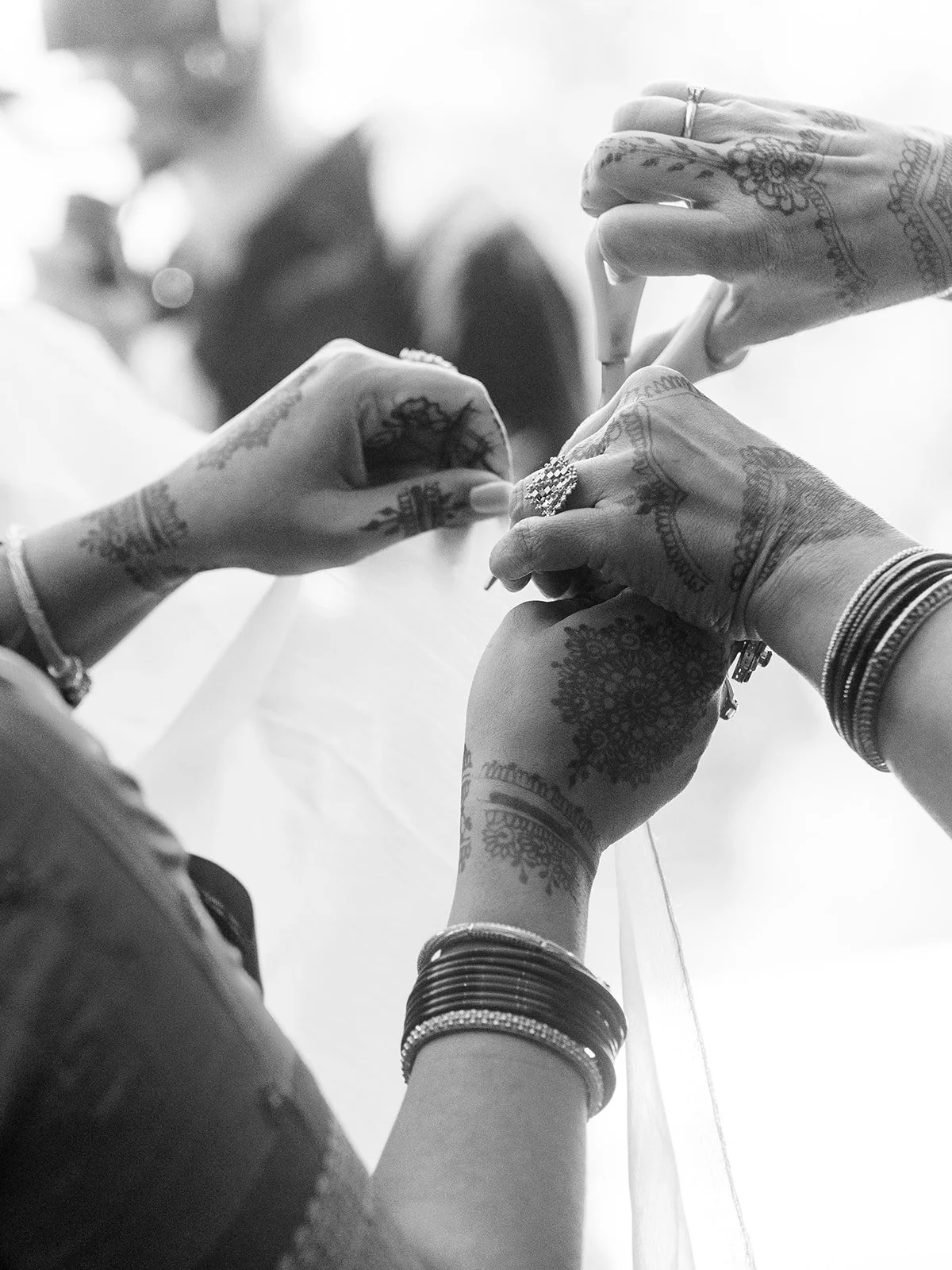 Black and white close-up of henna-adorned hands and bangles fastening a veil during bridal preparations.