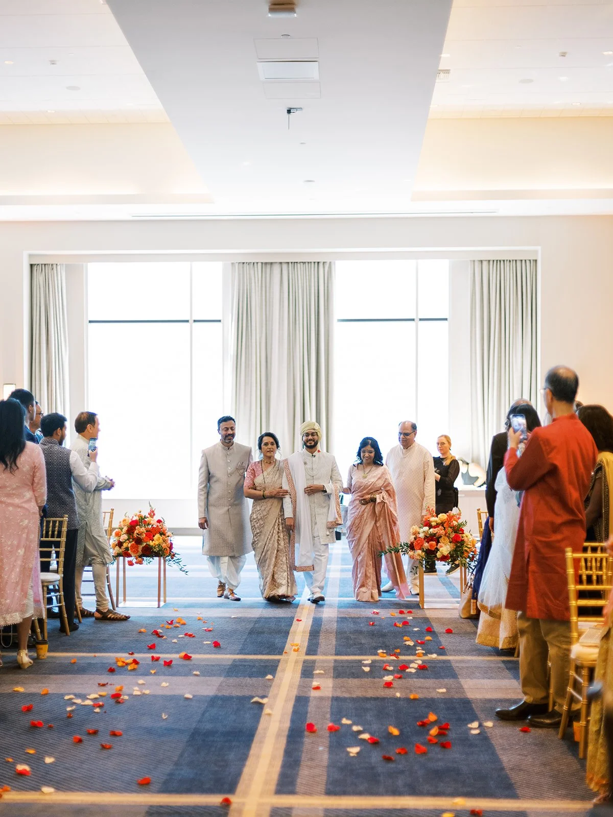 Groom in ivory sherwani and turban entering ceremony with family down petal-lined aisle.
