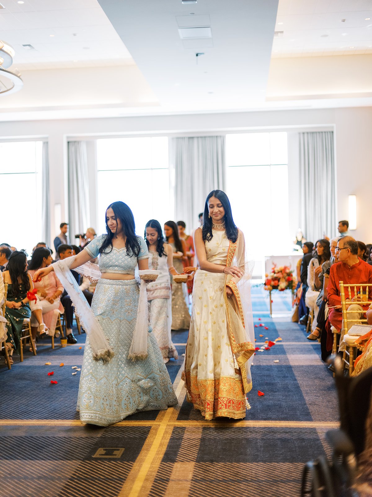 Bridesmaids in pastel lehengas walking down aisle scattering rose petals at Hindu wedding ceremony.