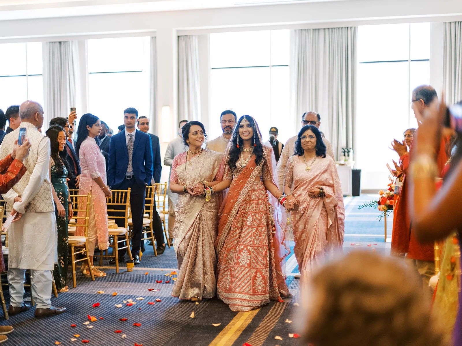 Bride in orange lehenga walking down aisle with family during traditional Hindu wedding ceremony.