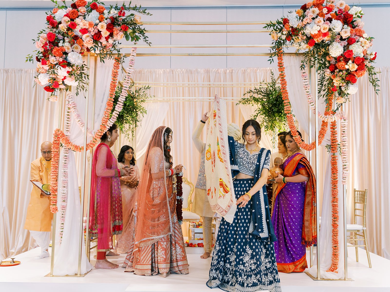 Couple standing beneath floral mandap with priests and family during Hindu wedding ceremony.