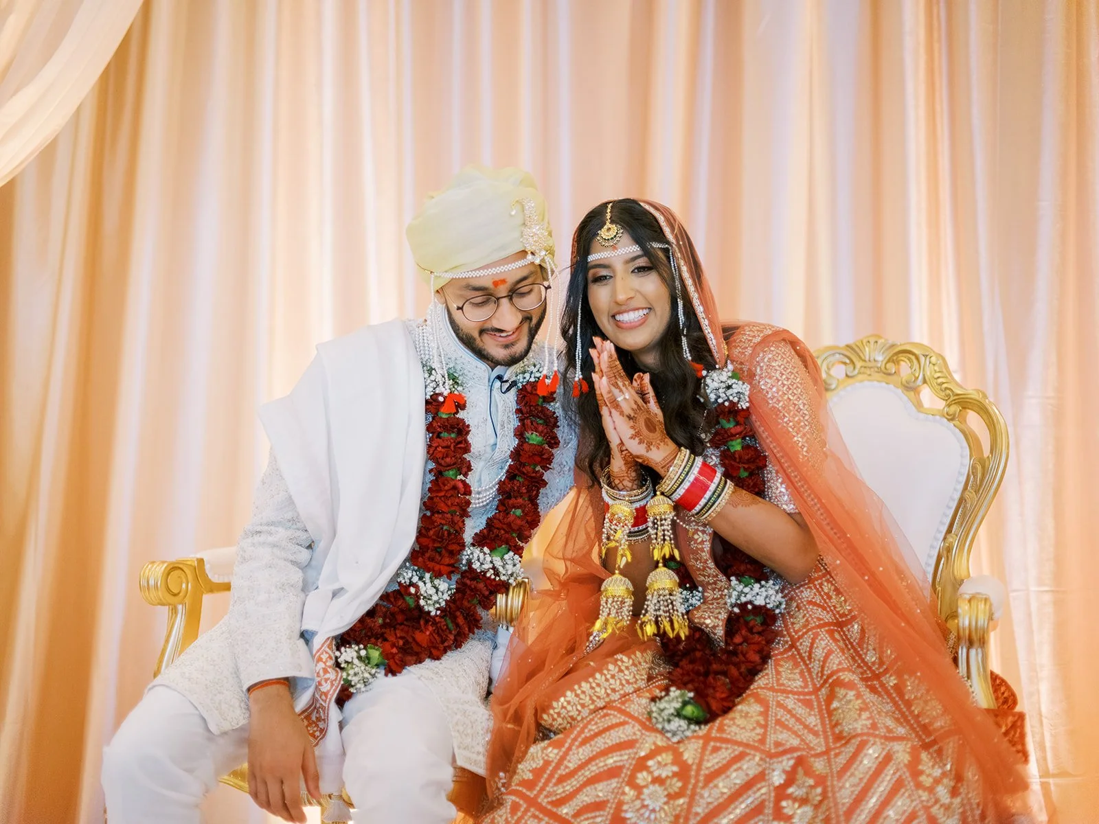 Bride and groom seated together under mandap, smiling in orange lehenga and ivory sherwani with red garlands.