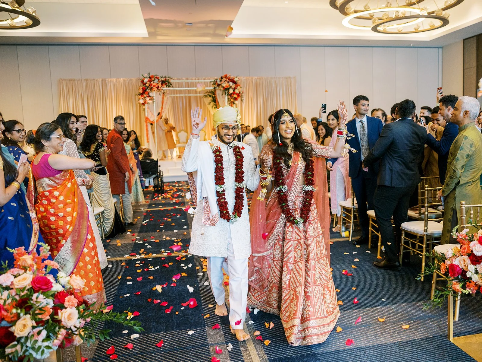 Bride and groom walk down petal-lined aisle smiling as guests celebrate after Hindu wedding ceremony.