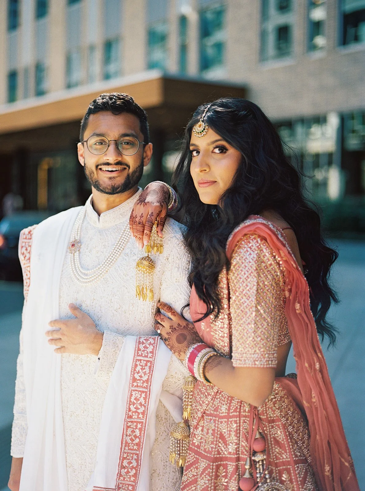 Bride resting henna-adorned hand on groom’s shoulder during outdoor Indian wedding portraits.
