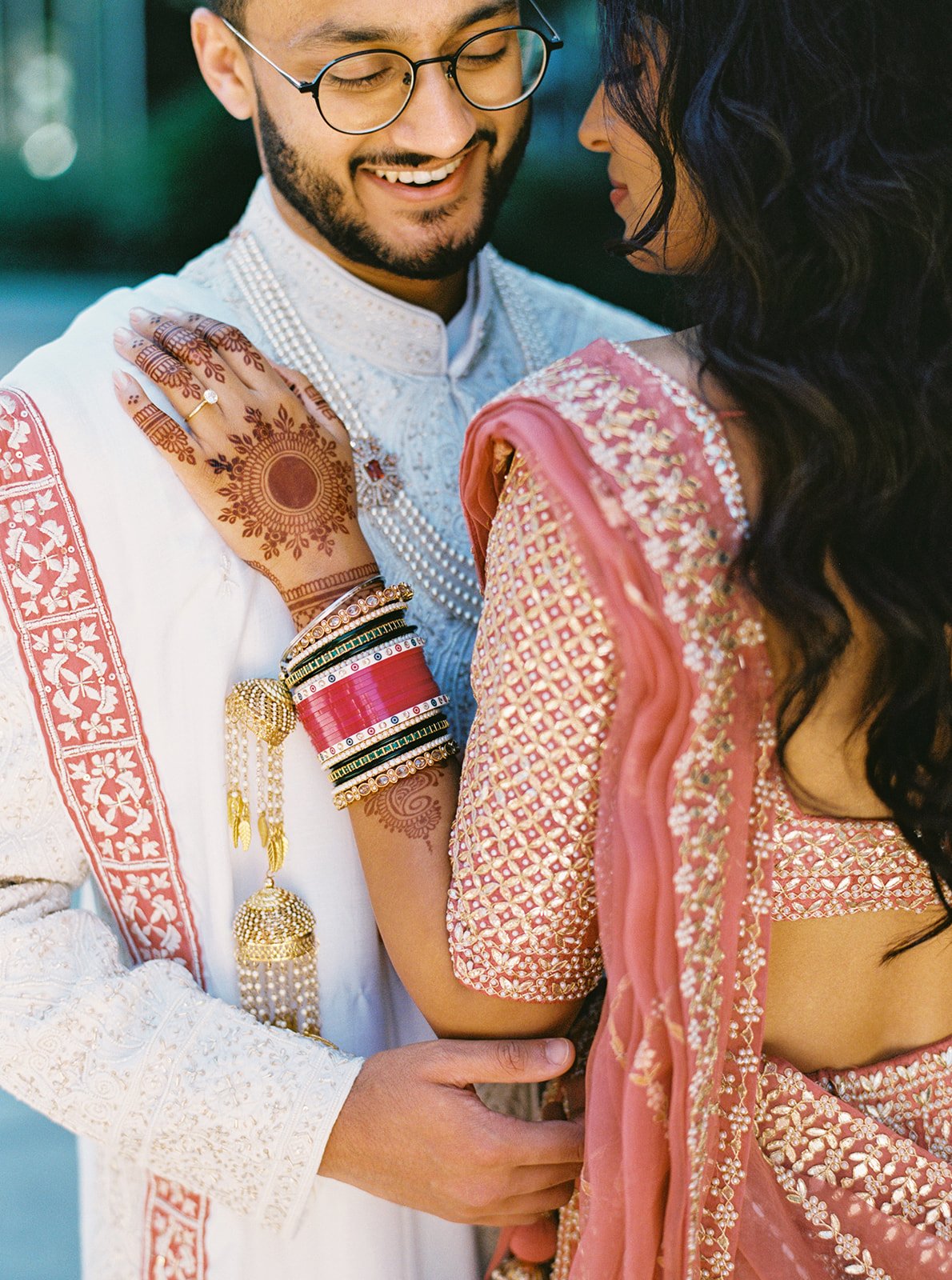 Close-up of bride’s mehndi hand and red chooda bangles resting on groom’s sherwani.