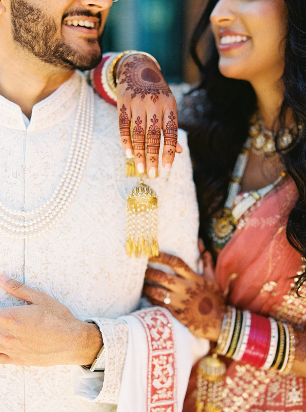 Close-up of bride’s henna hand with gold kalire jewelry against groom’s ivory outfit.