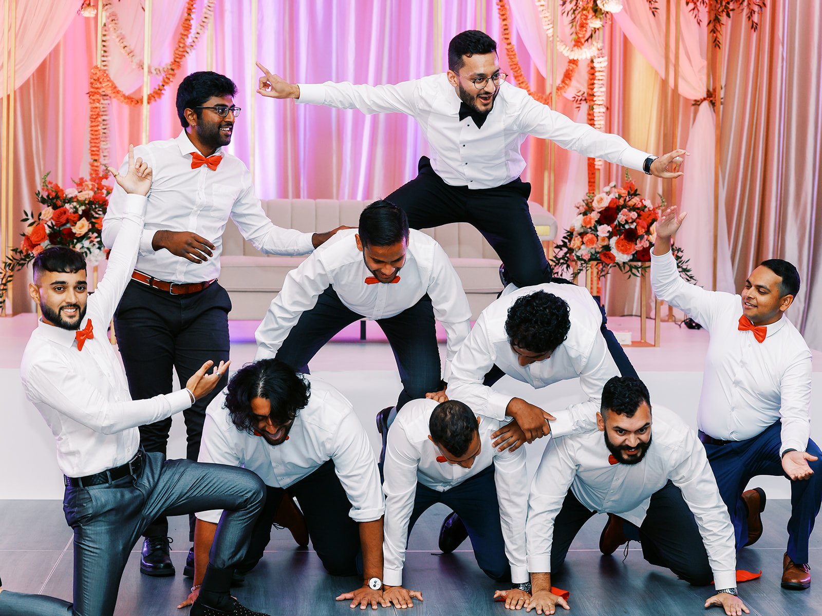 Groomsmen in white shirts and red bow ties forming pyramid during reception dance performance.
