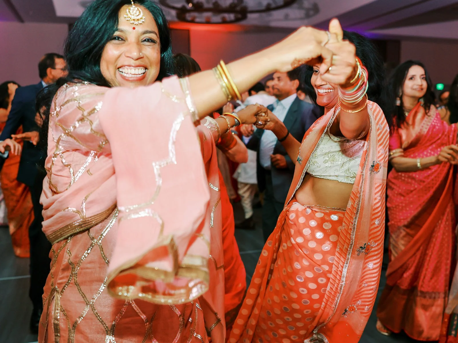 Two women in pink and orange sarees laughing and dancing together at a wedding reception.