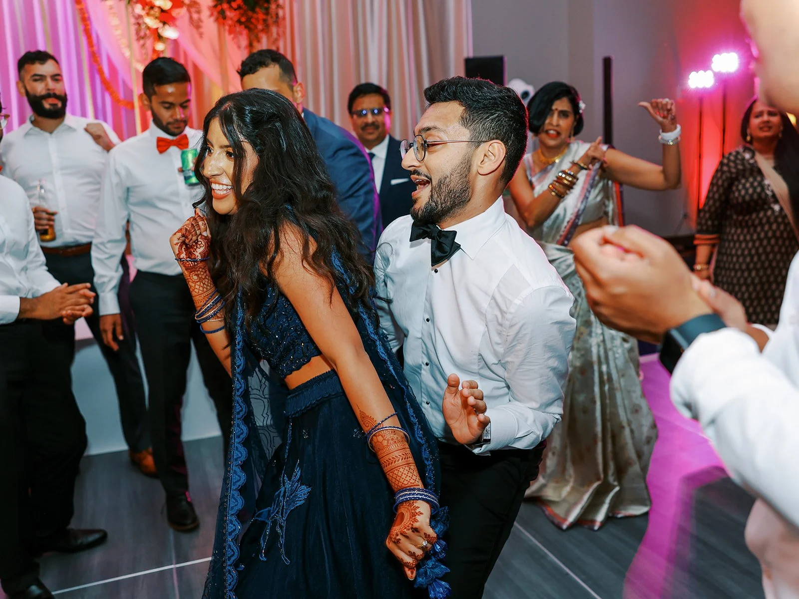 The bride in a navy lehenga and groom in a bow tie dancing and laughing together surrounded by celebrating guests.