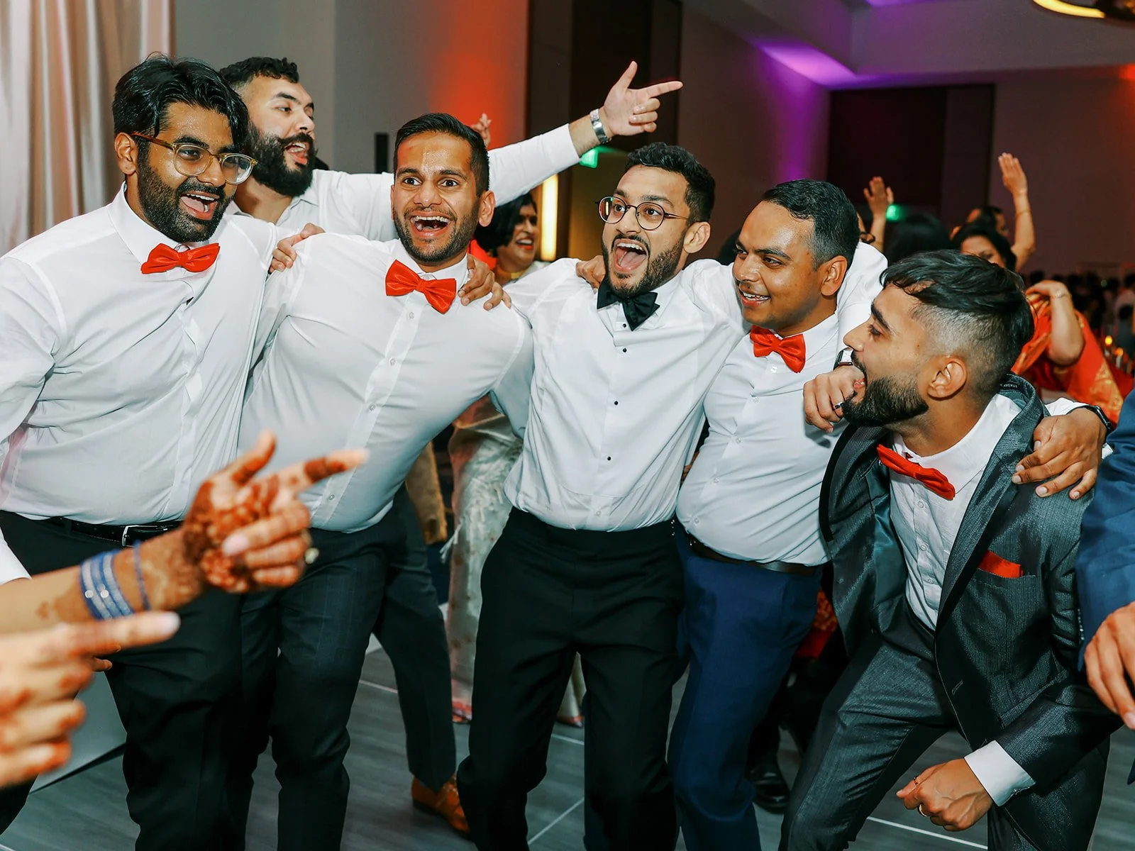 A group of groomsmen in white shirts and red bow ties cheering and dancing together at the reception.