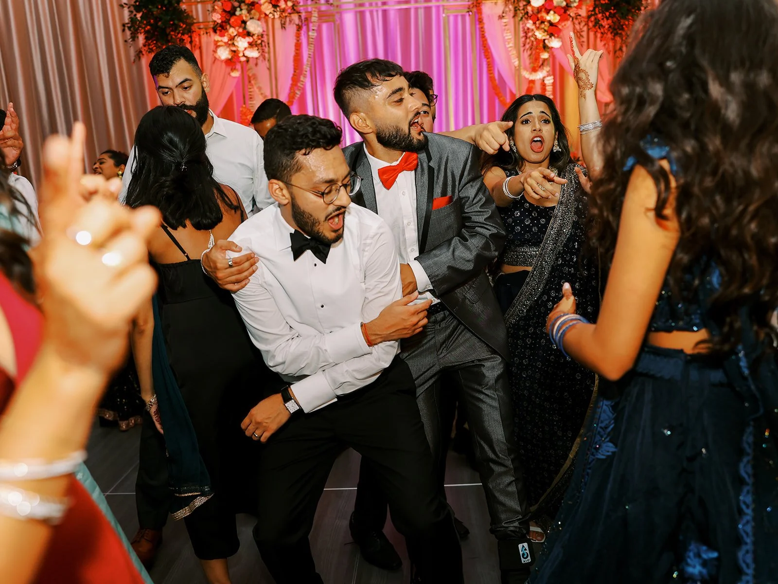 A group of energetic guests in suits and sarees dancing wildly on the wedding reception dance floor.