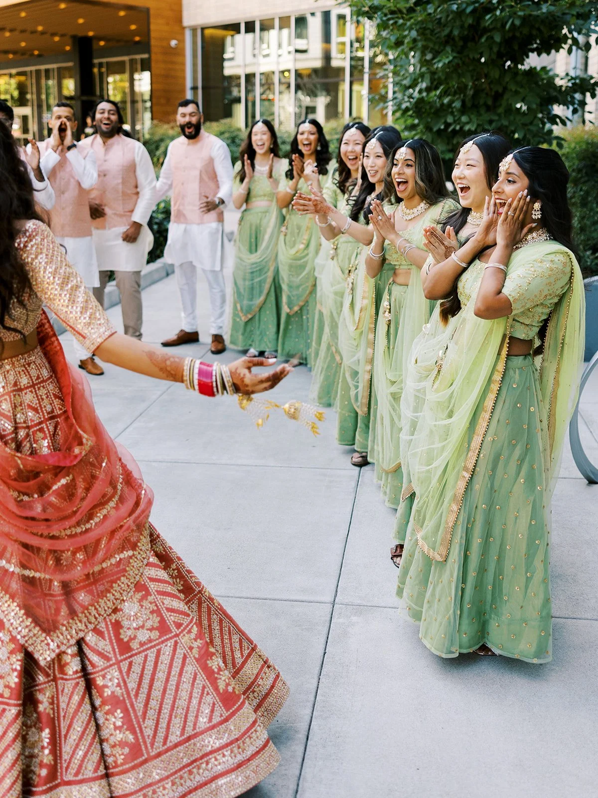 Bridesmaids in sage green lehengas clapping and reacting as bride spins in red bridal outfit.