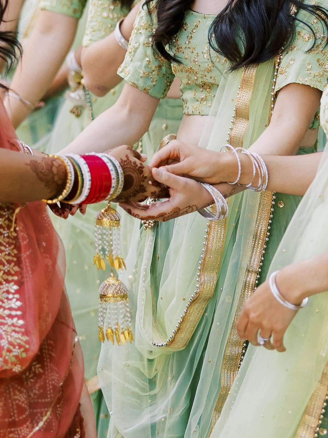 Close-up of bride’s mehndi hands with red chooda bangles as bridesmaids hold her hands.