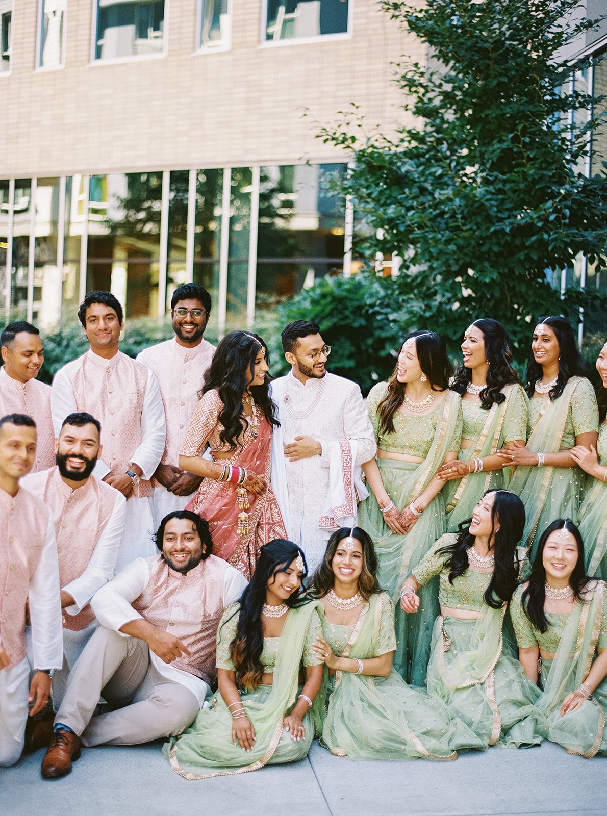 Bride and groom laughing with bridal party in coordinated green and blush outfits.