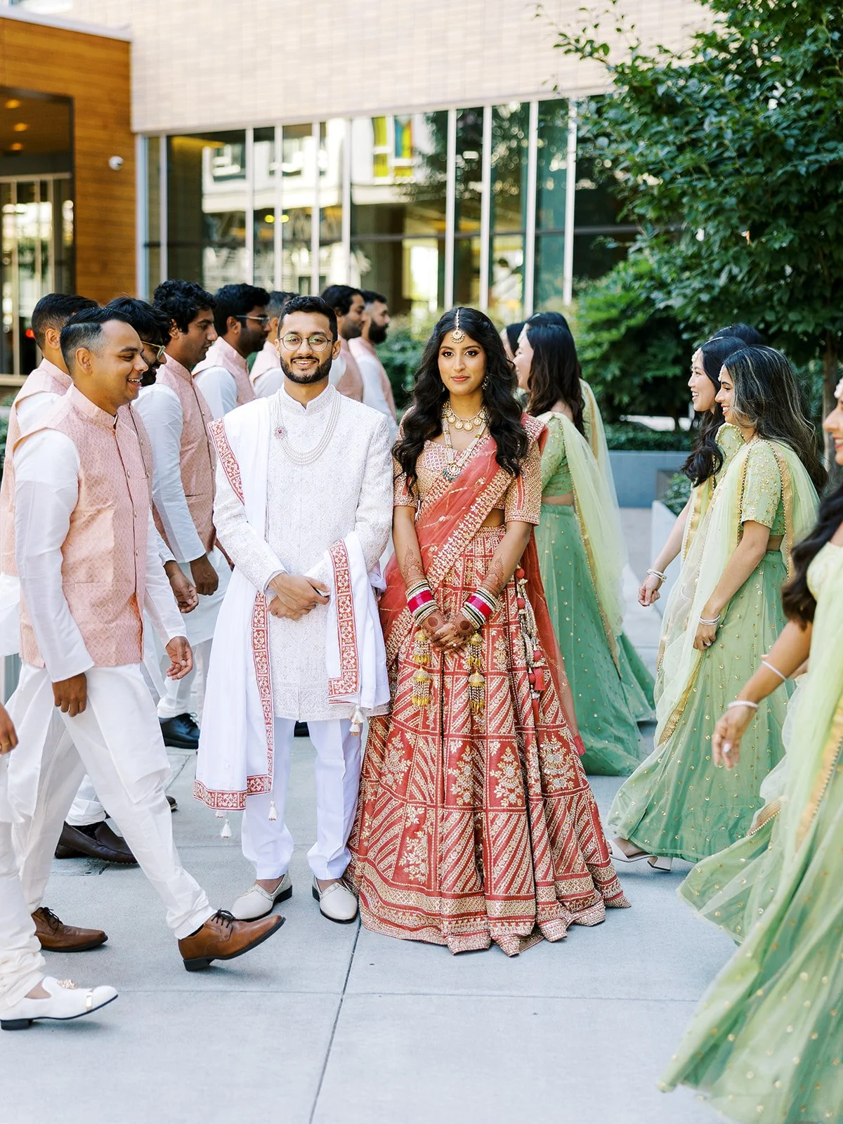 Bride and groom standing together as bridal party walks around them outdoors.