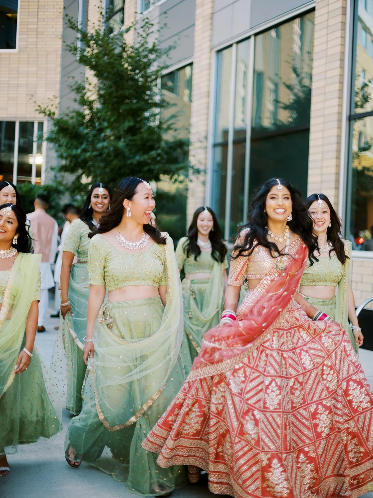 Bride walking forward in red and gold lehenga with bridesmaids in green lehengas behind her.