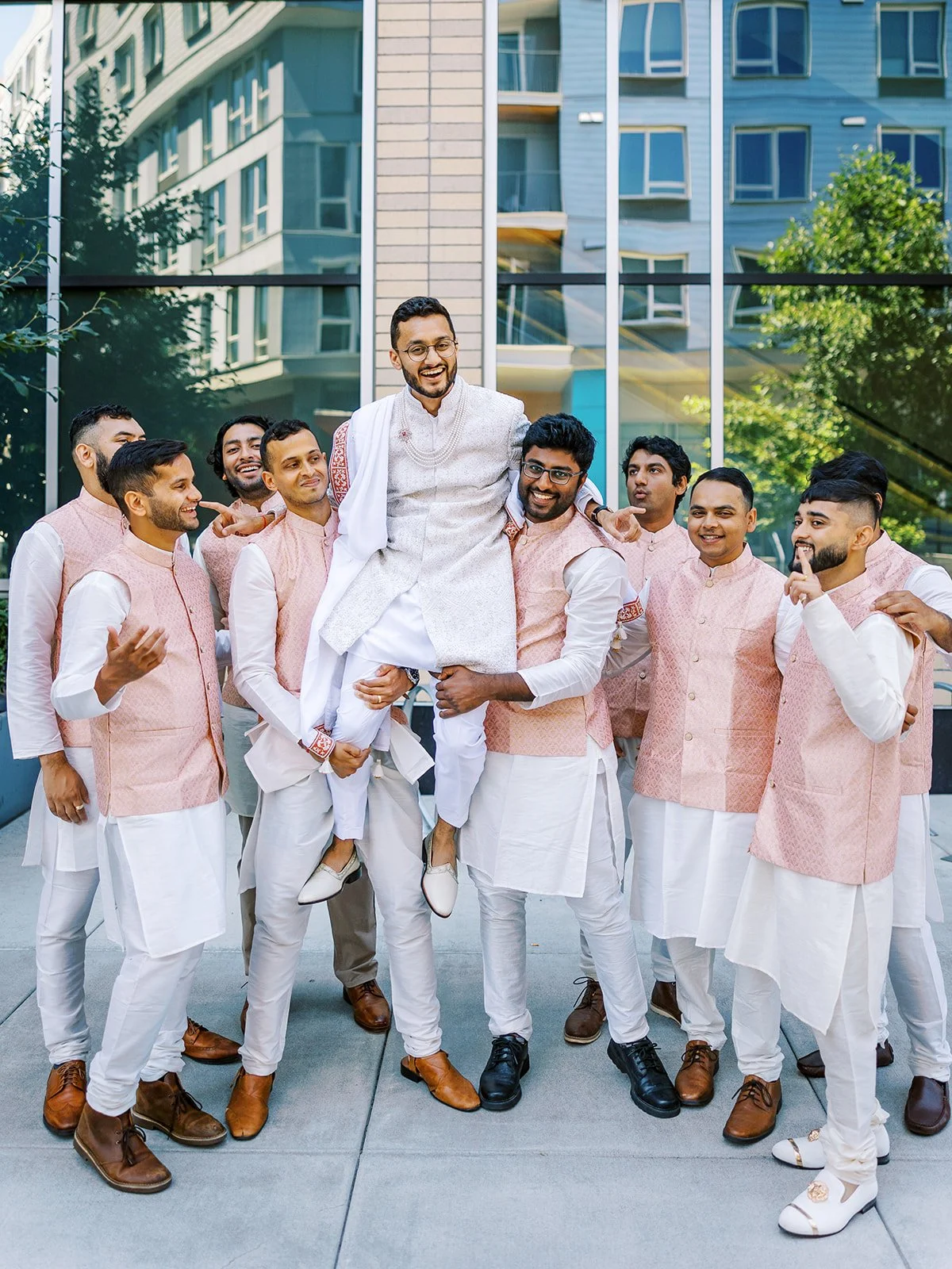 Groomsmen lifting groom in white sherwani during lively outdoor wedding portraits.