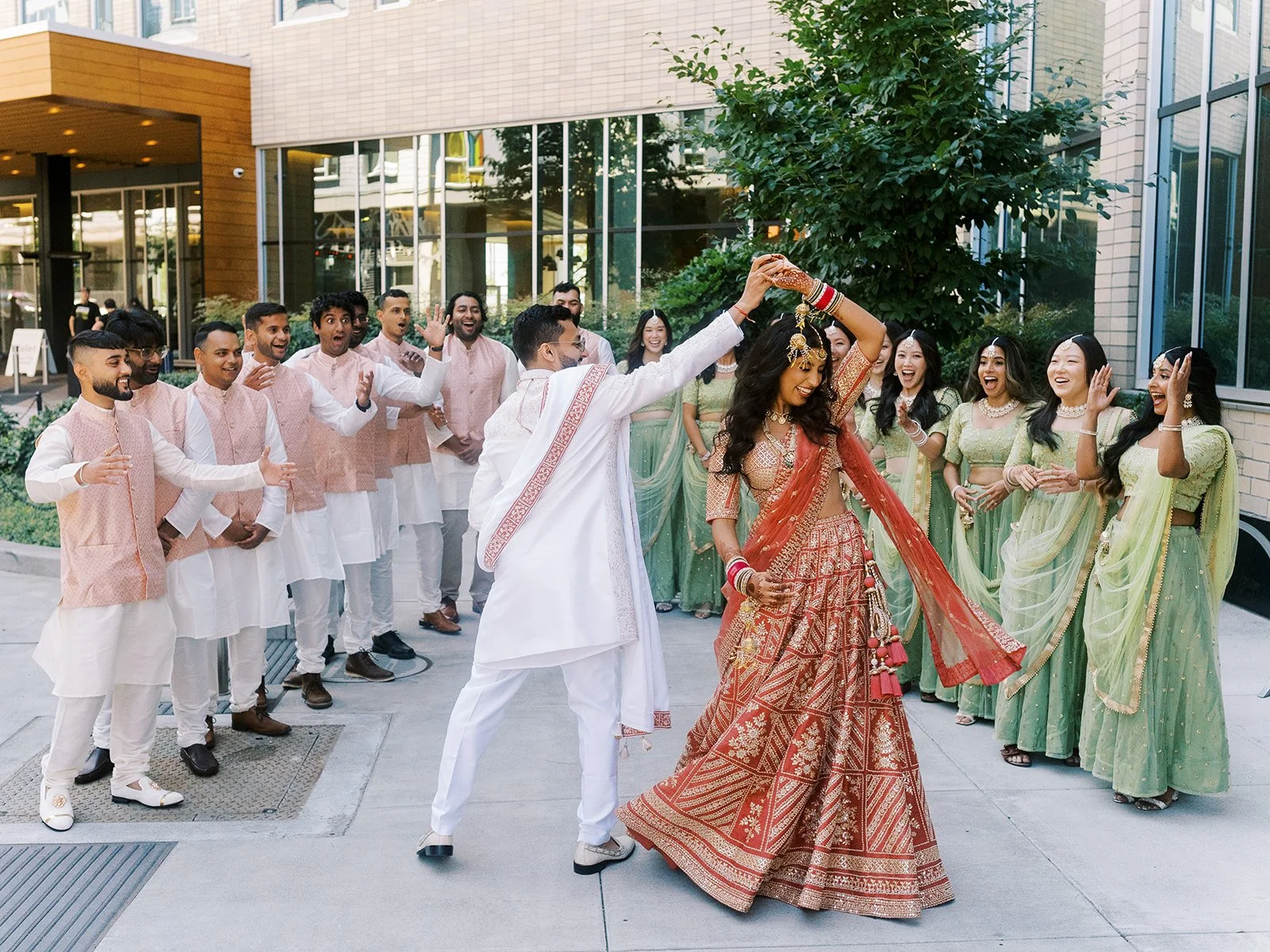 Bride in red and gold lehenga twirling as groom and bridal party cheer outside modern venue.