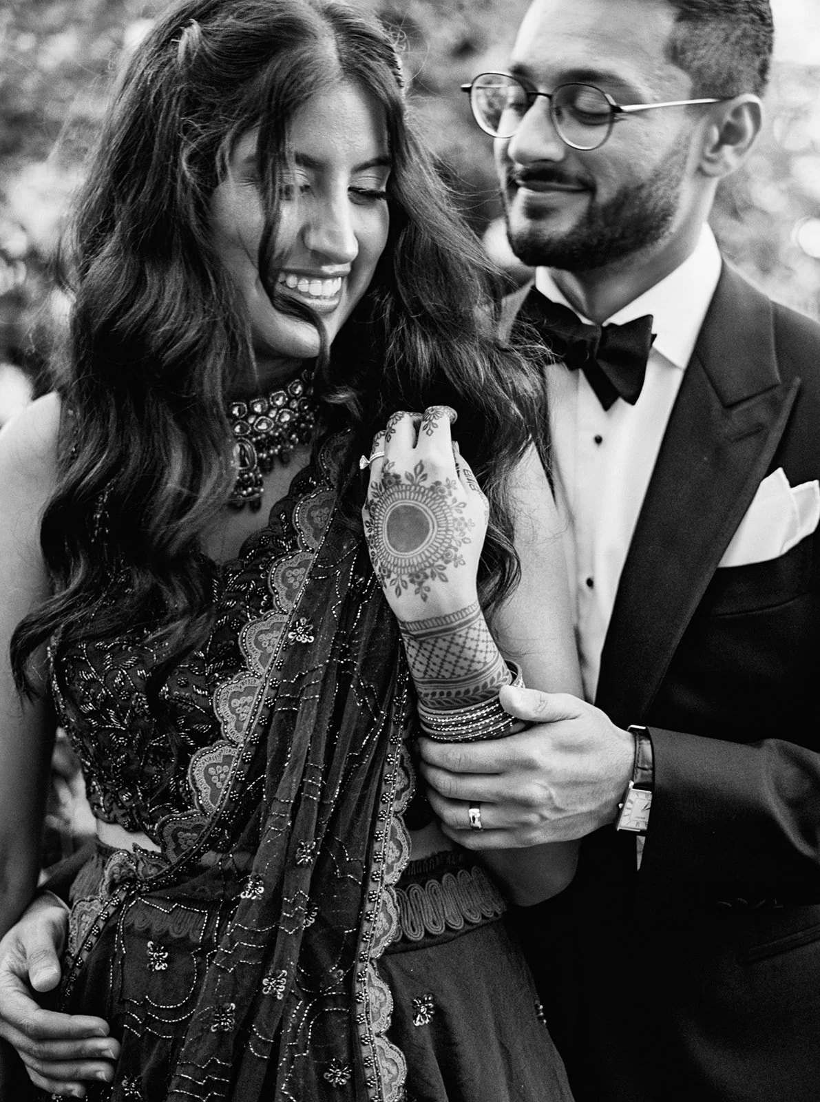 Black and white close-up of a laughing bride in an embroidered saree with mehndi, embraced by a groom in a tuxedo.