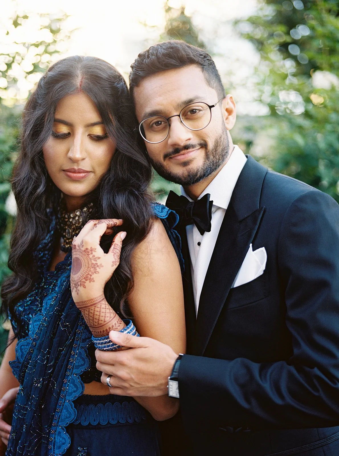 A groom in a tuxedo holds a bride close from behind as she looks down, her mehndi-adorned hand resting on his arm.