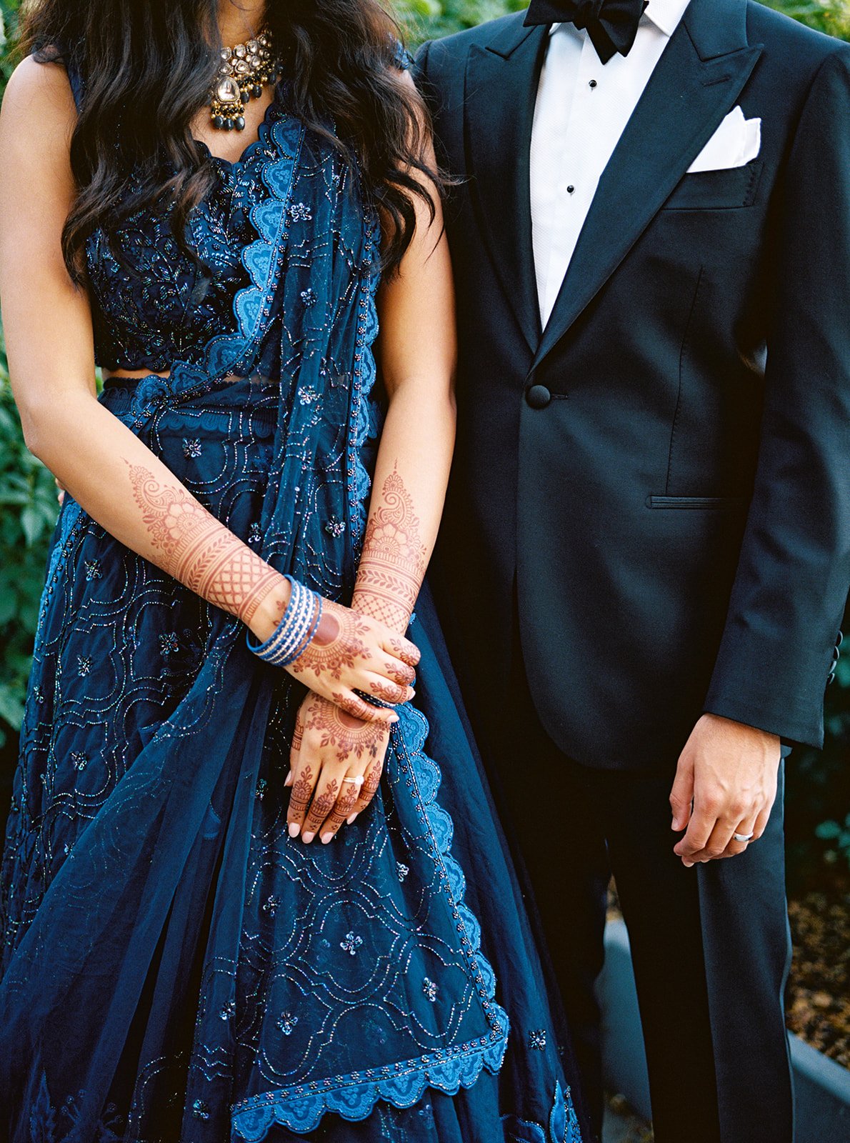  Close-up of a bride's mehndi-covered arms and blue bangles against her navy lehenga, beside a groom in a tuxedo.