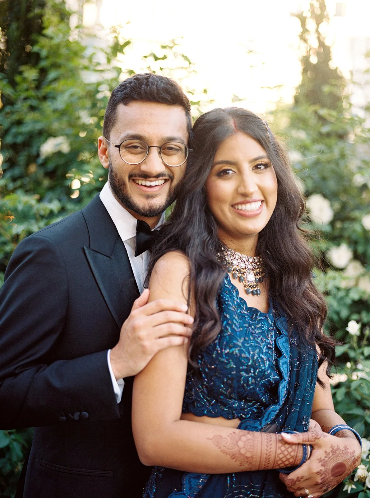 A smiling couple outdoors — groom in a tuxedo embracing a bride in a blue embroidered lehenga with mehndi and statement jewelry.