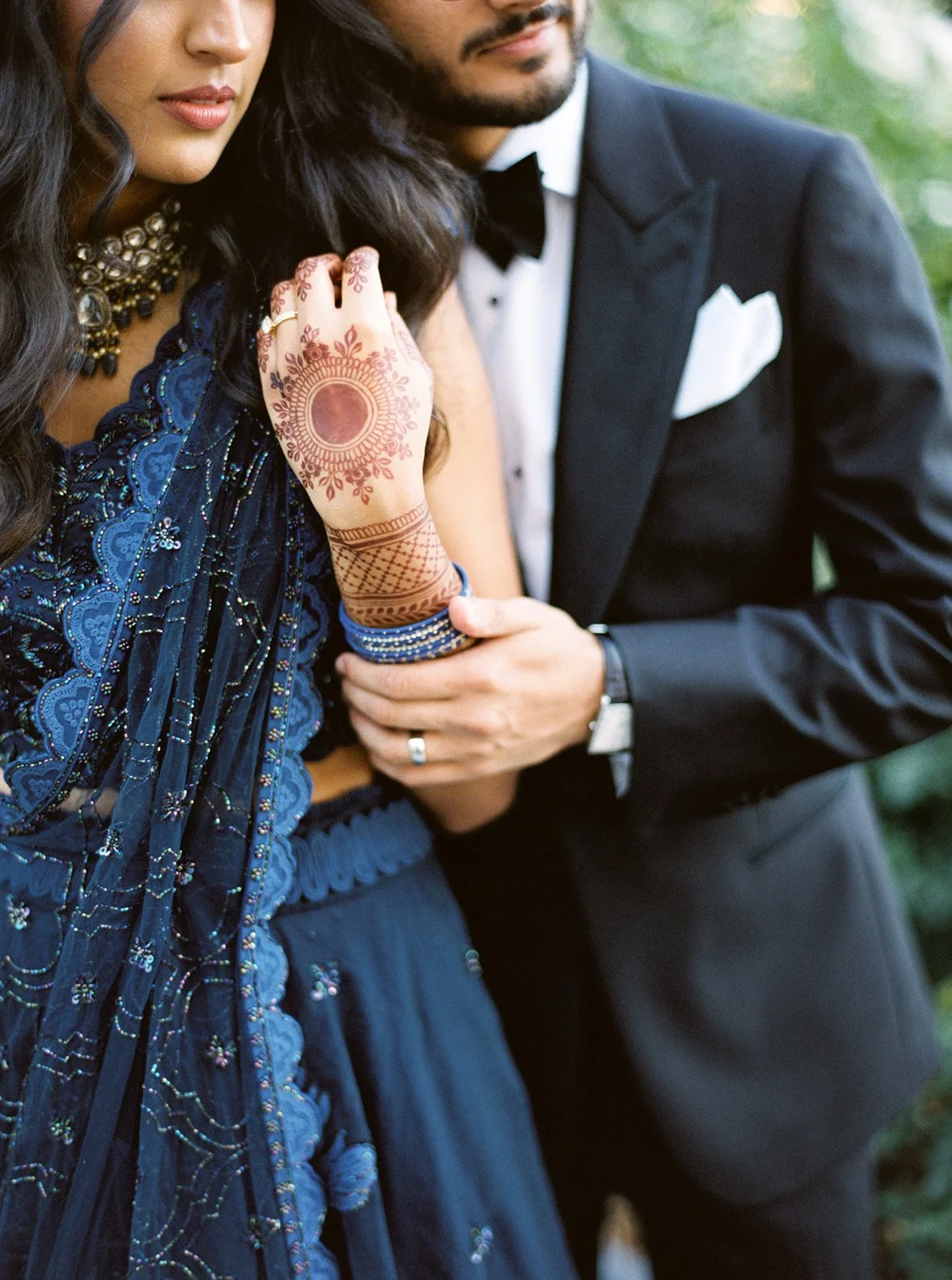  Close-up of a bride's intricately hennaed hand and blue bangles against her navy lehenga, held gently by a groom in a tuxedo.
