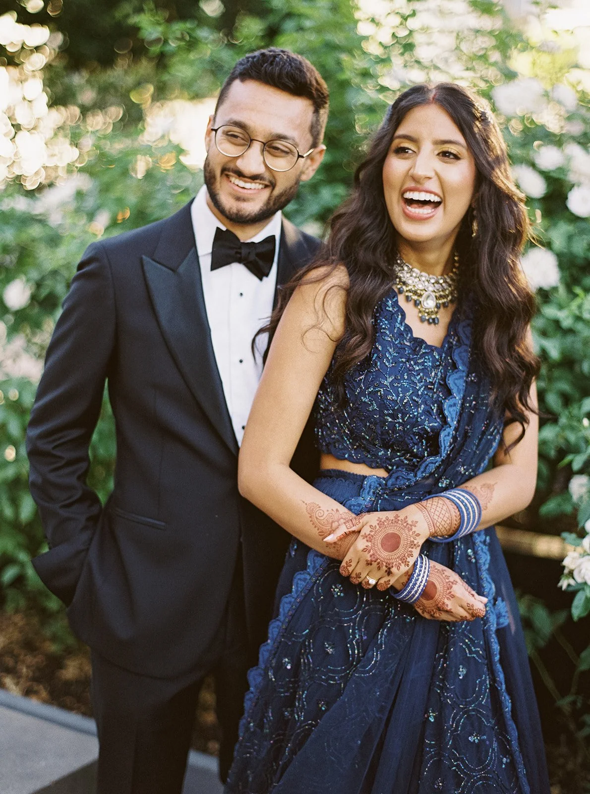 A joyful couple laughing outdoors — groom in a tuxedo, bride in a blue lehenga with mehndi and bangles.