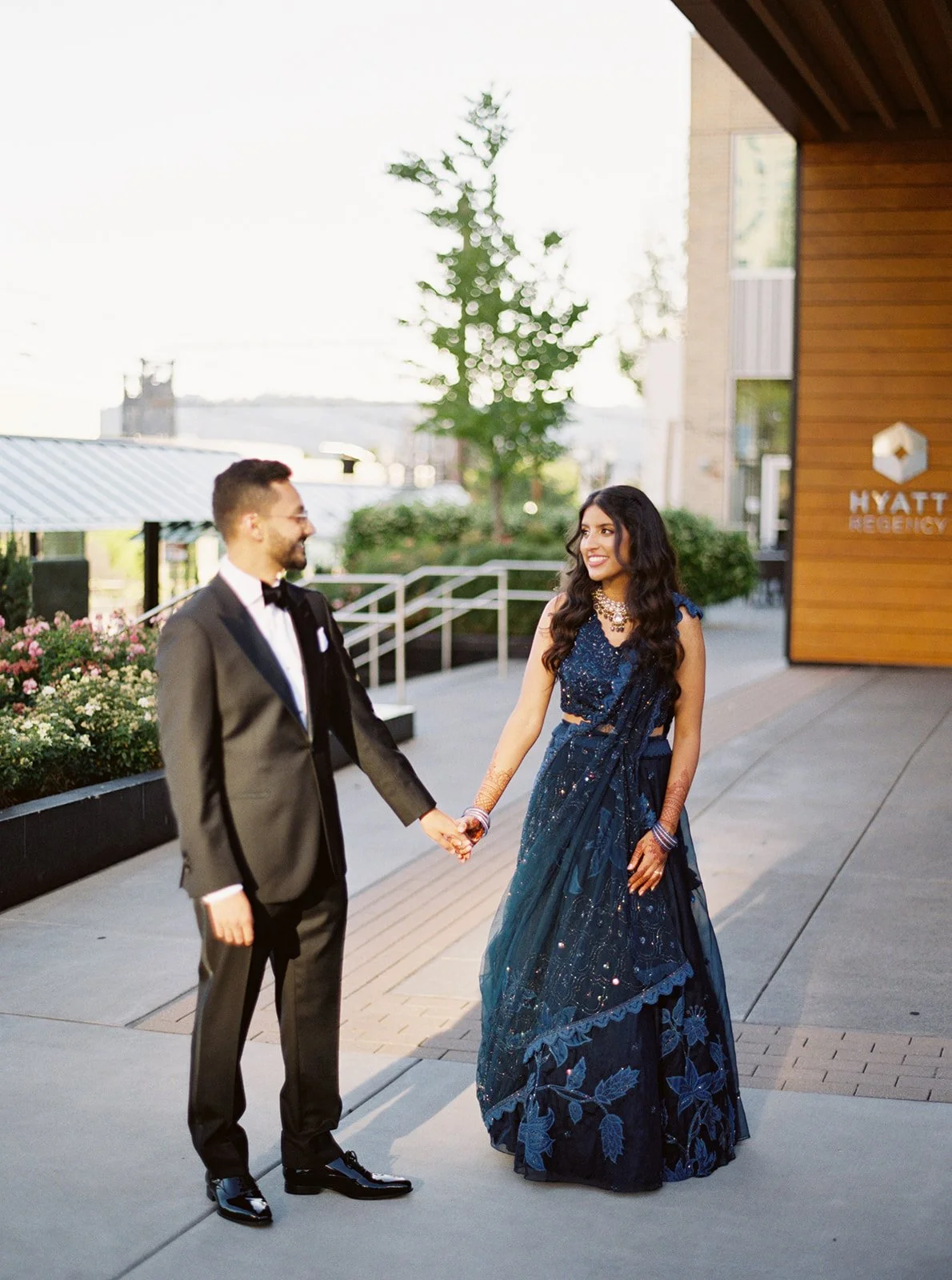A couple holding hands outside the Hyatt Regency — groom in a tuxedo, bride in a navy embroidered lehenga with mehndi.