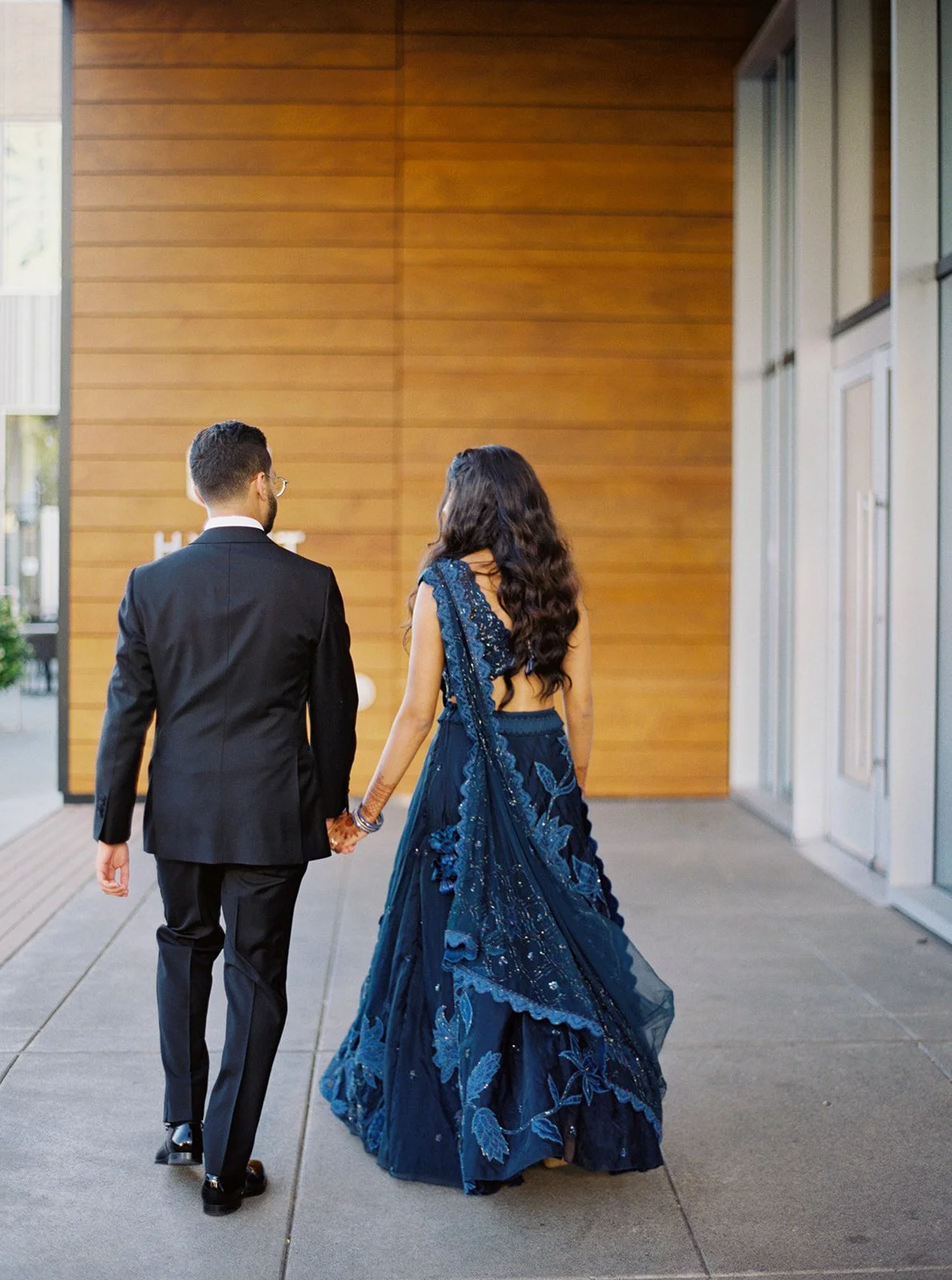 A couple walking away hand-in-hand outside the Hyatt Regency, the bride's navy lehenga trailing behind her.
