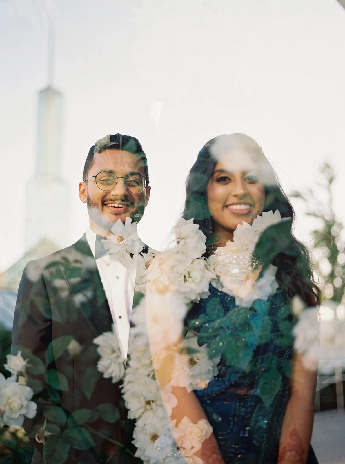 A dreamy double-exposure portrait of a smiling couple in wedding attire layered with white roses and greenery.