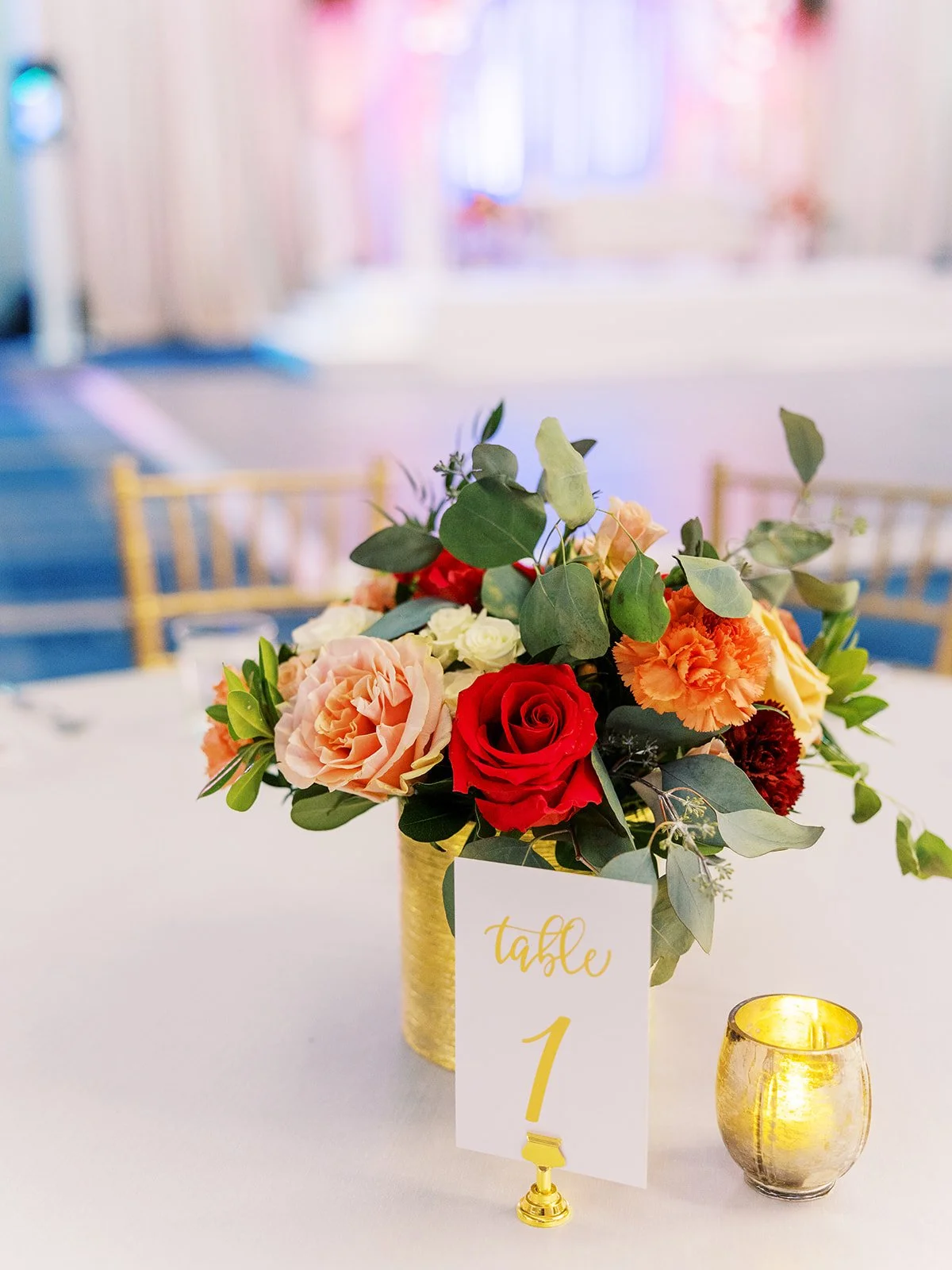 Gold vase centerpiece with red and peach roses, greenery, and table number card at reception.