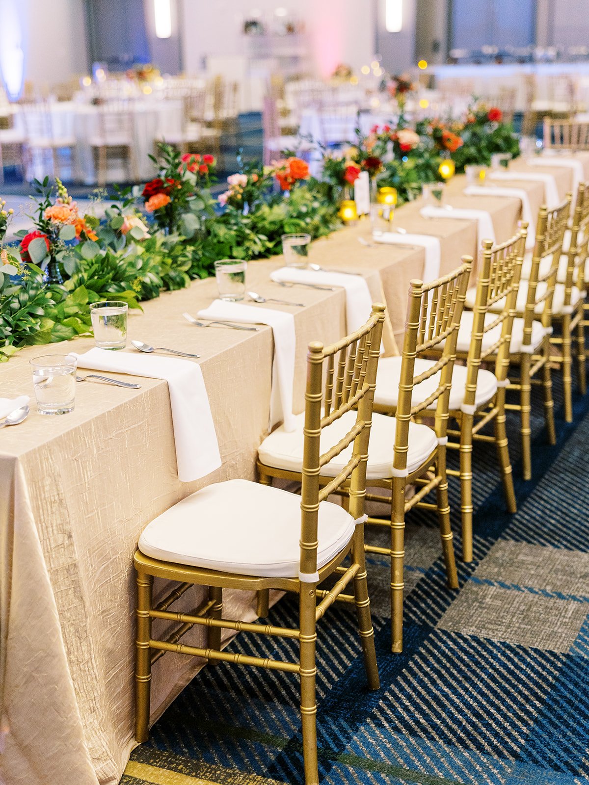 Gold vase centerpiece with red and peach roses, greenery, and table number card at reception.Wedding reception head table with gold chiavari chairs and lush floral garland centerpiece.
