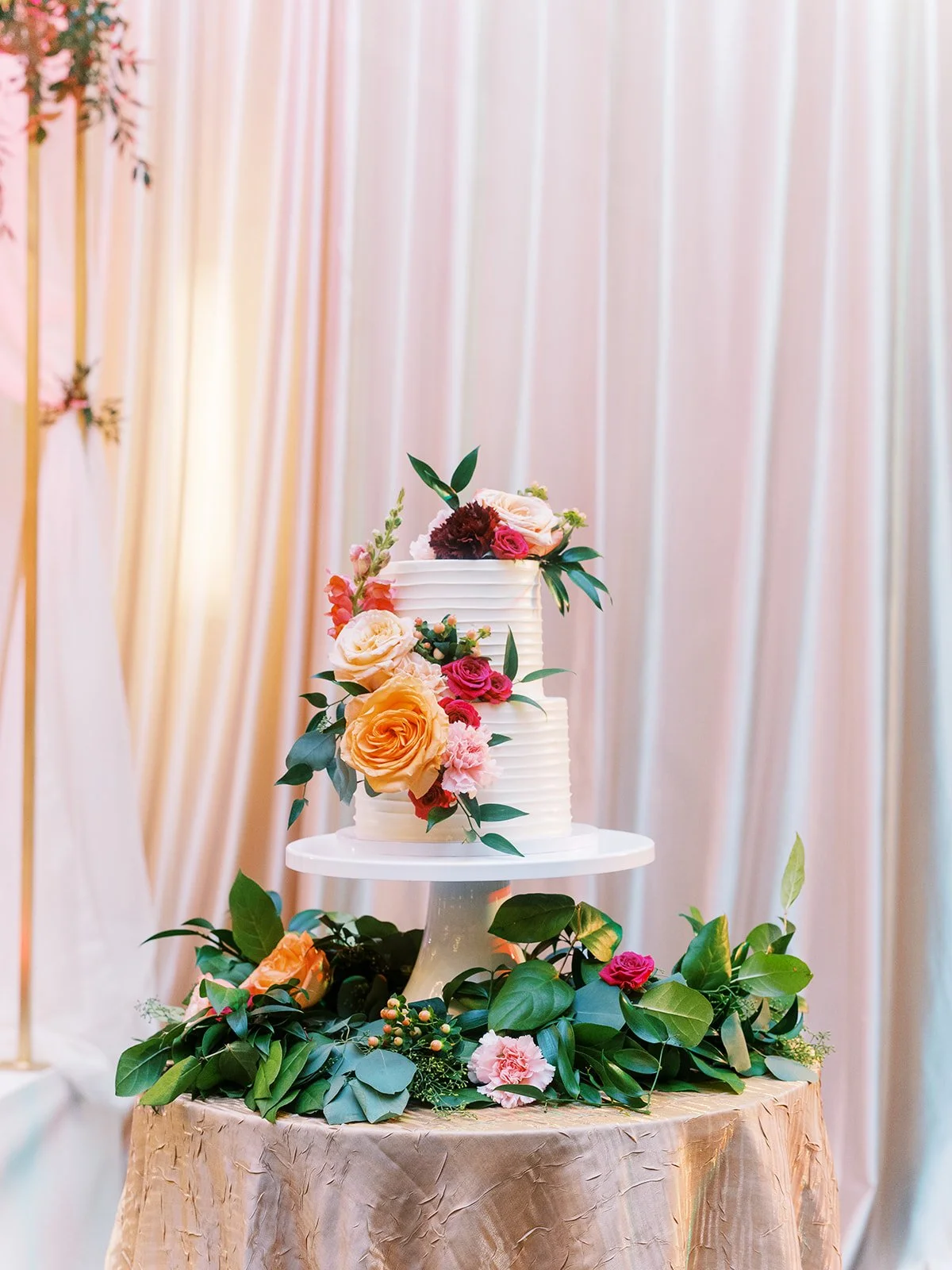 White two-tier wedding cake decorated with peach, red, and pink flowers on gold linen table.