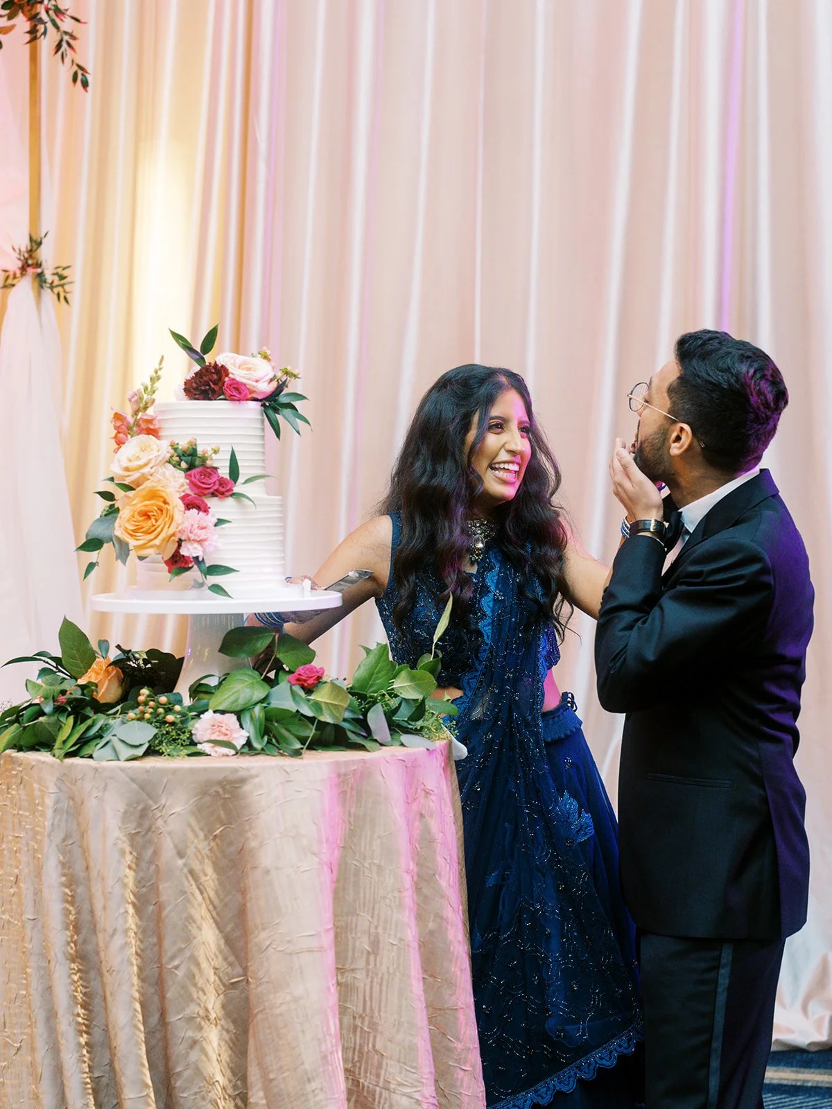 Couple in navy gown and black tuxedo cutting floral wedding cake at reception.