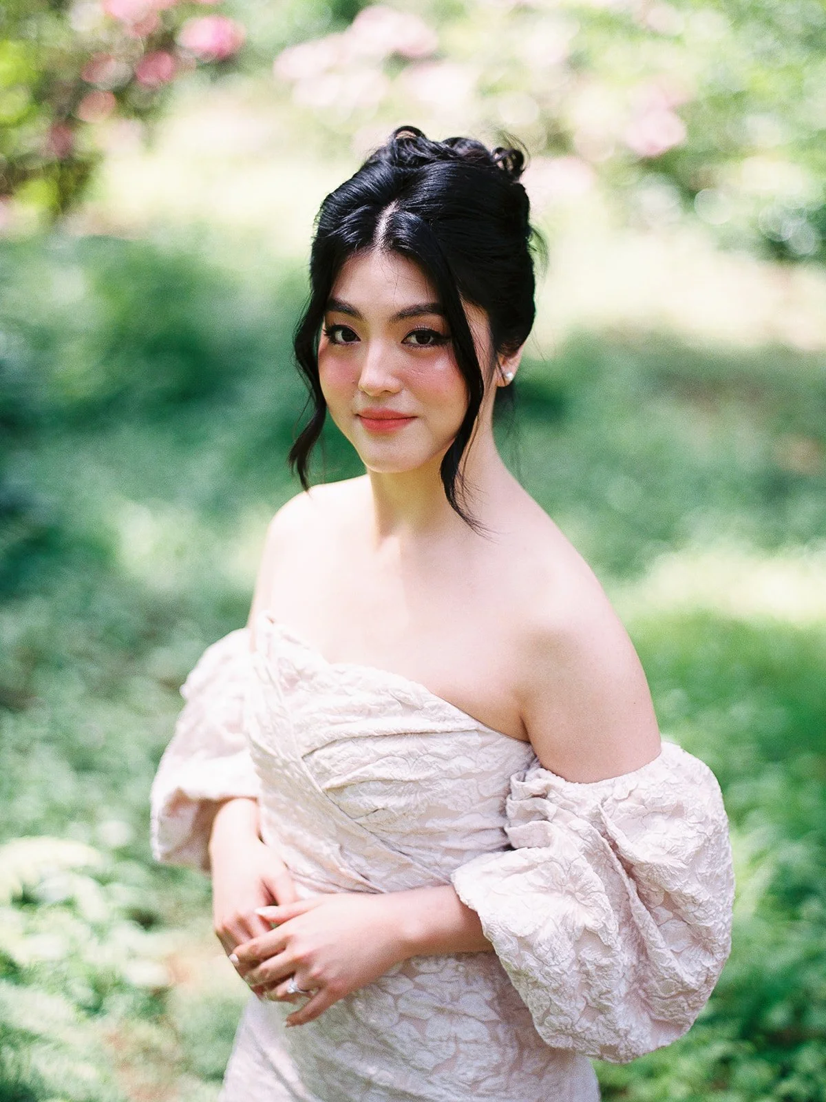 Portrait of bride with dark updo and off-shoulder dress standing in a garden with soft green foliage.