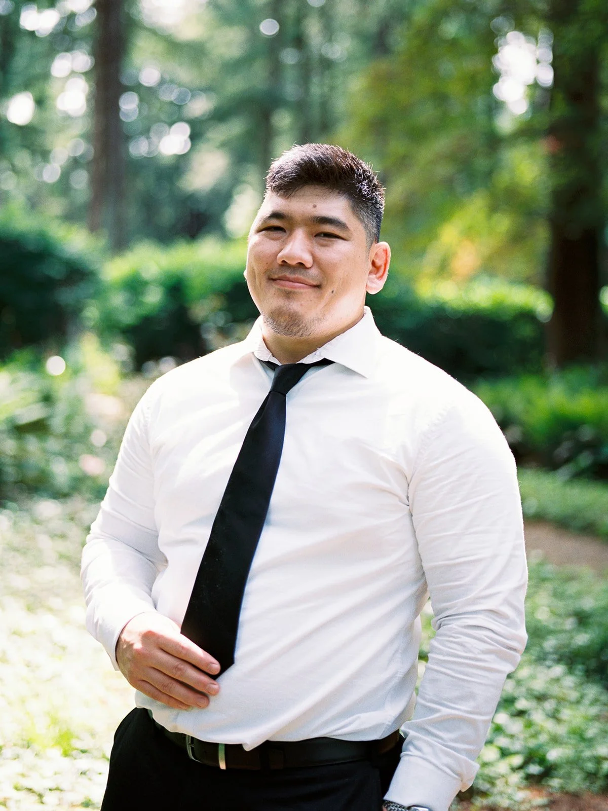 Groom standing on a shaded garden path wearing a white shirt and black tie.